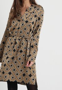 Langärmliges Kleid mit V-Ausschnitt, braunem und schwarzem geometrischem Muster, tailliertem Bund und seitlichen Schlitzen für mehr Bewegungsfreiheit. Glatte Stoffoberfläche.