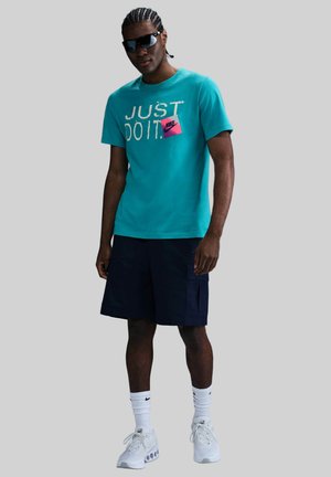 Camisa de manga corta turquesa con el texto "JUST DO IT" y logo de Nike rosa, combinada con pantalones cortos azul marino y zapatillas blancas. Lentes de sol puestos.