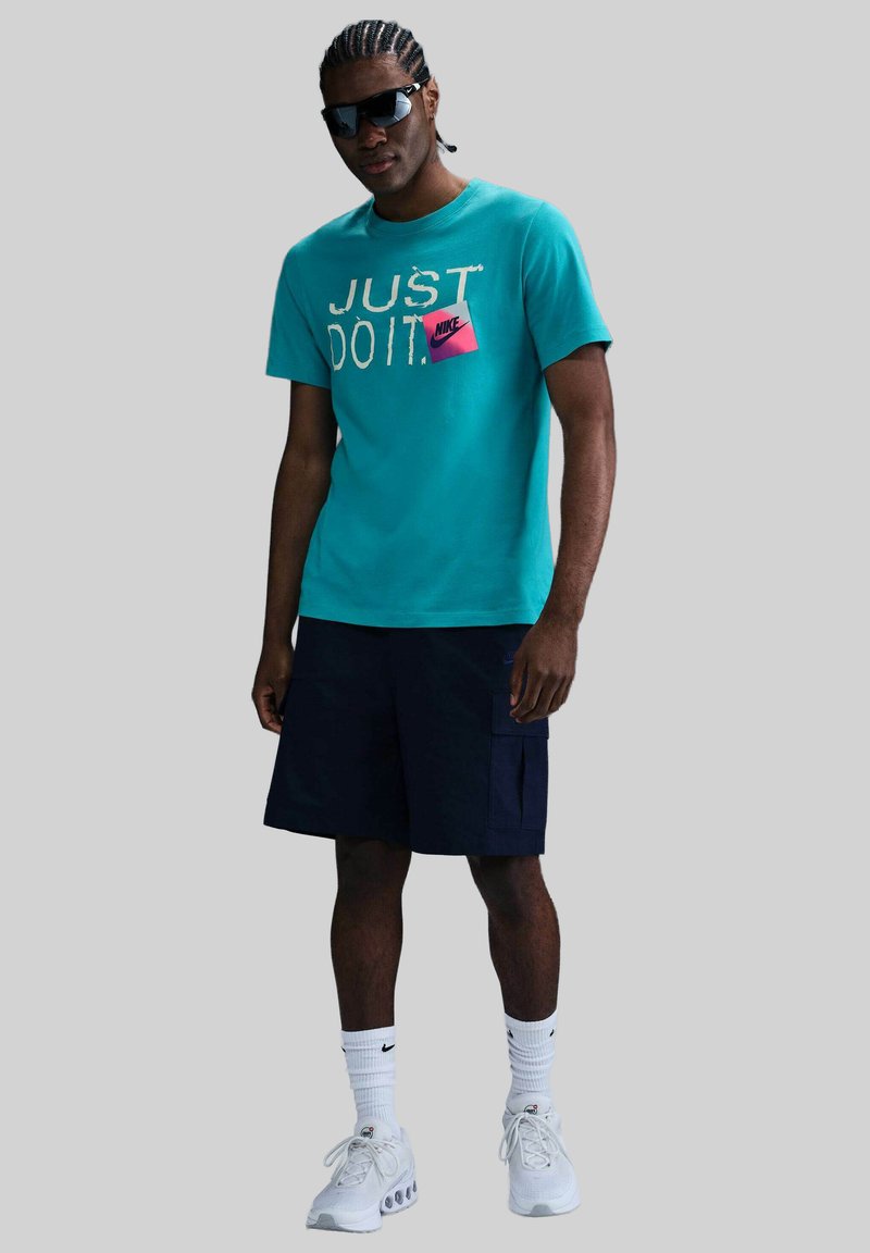 Camisa de manga corta turquesa con el texto "JUST DO IT" y logo de Nike rosa, combinada con pantalones cortos azul marino y zapatillas blancas. Lentes de sol puestos.