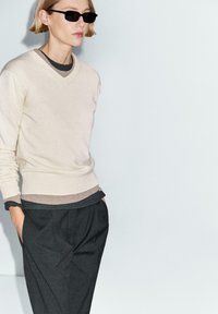 Person iført lag-på-lag beige og sort trøjer, sorte bukser og sorte solbriller, stående foran en ensartet lys baggrund.