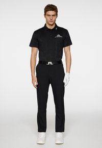 Black polo shirt with hexagonal texture, logo on the chest, paired with black trousers and a white belt. Model is wearing white sneakers and a glove.