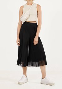 Zwarte geplooide culottes met een vloeiend ontwerp en een doorschijnende onderste laag, gecombineerd met een lichte crop top en witte sneakers, aangevuld met een klein wit tasje.