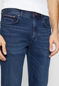 Temno modri traper jeans s klasičnim pet-pocket dizajnom, zlatimi dodatki in subtilno teksturirano površino.