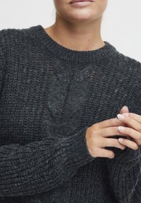 Donkergrijze gebreide trui met een ronde hals, textuurpatroon, geribde manchetten en zichtbare stiksels. Handen gevouwen voor zich.