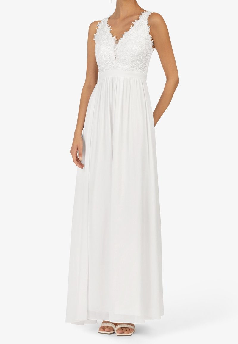 Robe maxi blanche sans manches avec corsage en dentelle fleurie, décolleté en V, taille cintrée et jupe fluide portée avec des talons blancs ouverts à l'avant.
