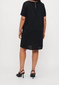 Vestido negro de manga corta con corte recto, que presenta una cremallera en la parte posterior. La tela parece suave y el dobladillo está por encima de la rodilla.