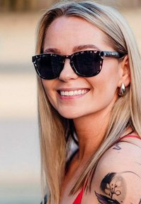 Černé sluneční brýle s vzorovaným rámem, doplněné dlouhými rovnými blond vlasy. Kulaté stříbrné náušnice a květinové tetování na ruce.