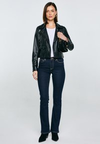 Žena u crnoj kožnoj jakni, bijeloj majici, tamnoplavim trapericama i crnim čizmama, drži malu crnu torbicu na neutralnoj pozadini.