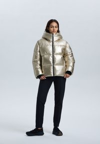 Metallisk guld puffjacka med hög krage och blixtlås, med en boxig design och ribbade svarta manschettdetaljer, tillsammans med svarta byxor.
