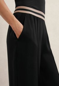 Mustad, lahtised püksid, millel on sile tekstuur, laia vöökohaga heledas beežis mustade triipudega ja küljetaskutega.