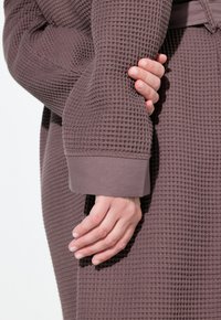 Robe en tissu mauve à texture gaufrée avec des manches à revers. Main reposant sur le bras, mettant en valeur le matériau doux et texturé.