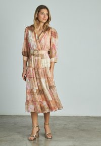 Blumengemustertes, geschichtetes Midi-Kleid in Pastellrosa, Gold- und Cremetönen. Mit V-Ausschnitt und tailliertem Gürtel. Kombiniert mit metallischen Blockabsatzsandalen.