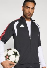 Fekete sportdzseki piros részletekkel és fehér panelekkel, sima anyagból. A modell egy mintás focilabdát tart.