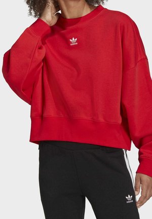 Osoba ubrana w czerwoną bluzę Adidas i czarne spodnie Adidas z logo, stojąca na jednolitym białym tle.