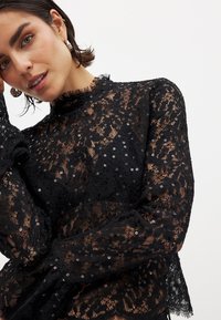 Schwarze Spitzenbluse mit floralen Mustern und Pailletten. Hoher Kragen, lange Ärmel mit Rüschensaum und strukturiertem Finish.