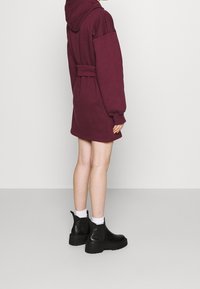 Bordeauxfarbener Kapuzenpullover-Kleid mit langen Ärmeln, Taillenkordelzug und seitlichen Taschen, kombiniert mit schwarzen Knöchelstiefeln und weißen Socken.