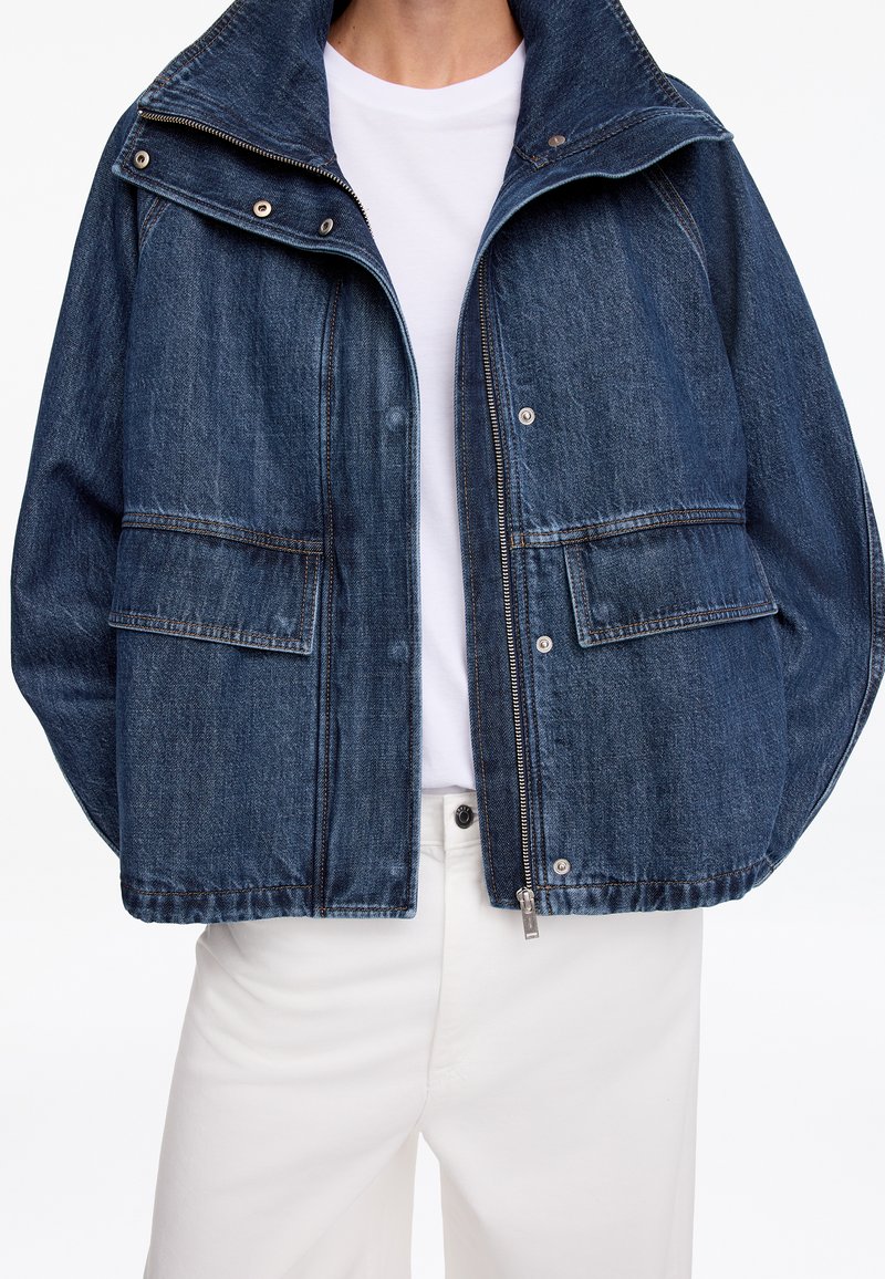 Personne portant une veste en jean bleu foncé avec poches avant sur une chemise blanche et un pantalon blanc, montrée du cou aux hanches.