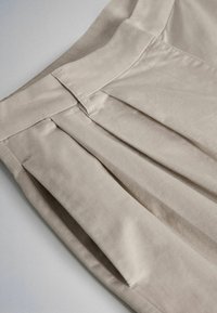 Tessuto beige plissettato con vita e passante per cintura, mostrando cuciture dettagliate e la texture del capo.