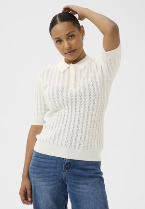 Kobieta o ciemnych włosach ubrana w białą, krótki rękaw, dzianinową koszulkę polo i niebieskie dżinsy, pozująca z jedną ręką uniesioną za głową.