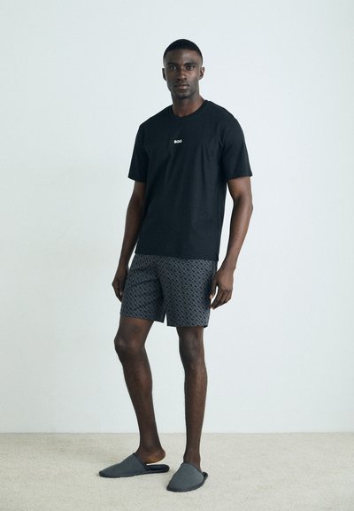 T-shirt noire avec logo blanc, shorts sombres à motifs et chaussures gris à enfiler. Tissu lisse, coupe décontractée, debout sur un tapis clair.