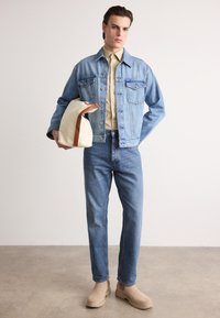 Denim jacket in light blue with two chest pockets, beige shirt underneath, blue jeans, and beige boots. Model holds a cream bag with brown strap.