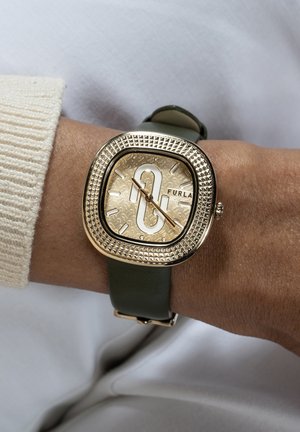 Orologio quadrato color oro con cassa texturizzata e cinturino in pelle verde. Presenta un quadrante dorato con numeri in rilievo e un logo.