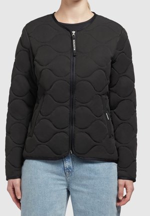 Persoon gekleed in een zwart doorgestikt jack met ritssluiting en golfpatronen, gecombineerd met lichtblauwe spijkerbroek, staand tegen een effen achtergrond.