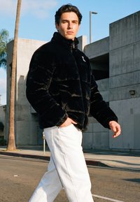 Muškarac u crnoj podstavljnoj jakni i bijelim hlačama hoda sunčanom ulicom blizu betonskih zgrada i palme.