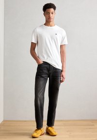 Hvit t-skjorte med rund hals og liten logo, kombinert med mørk denim jeans og gule sneakers. Modellen står på et trefloer mot en ensfarget vegg.