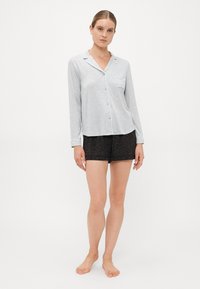 Kvinna som bär en ljusgrå långärmad skjorta med krage och svarta shorts med leopardmönster, stående barfota mot en vit bakgrund.