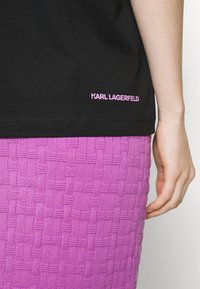 Primer plano de una mano al lado de una camiseta negra con el texto rosa "KARL LAGERFELD" encima de una falda texturizada morada.