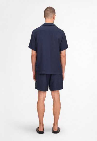 Homme debout de dos, portant une chemise bleu marine à manches courtes boutonnée, un short assorti et des sandales noires à enfiler sur fond blanc.