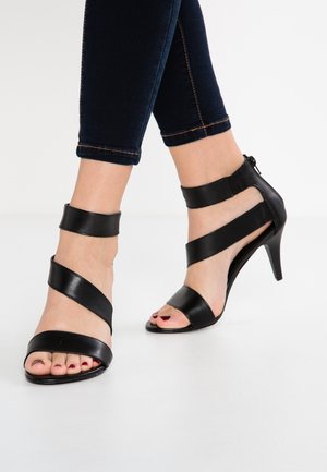 Zwarte hoge hakken sandalen hebben kruislings bij de enkels, een open teenontwerp en een glanzende afwerking, gecombineerd met donkere denim broeken.