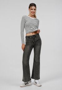 Black striped long-sleeve top, cropped design, paired with high-waisted grey flared jeans. Model wearing white sneakers with black stripes.