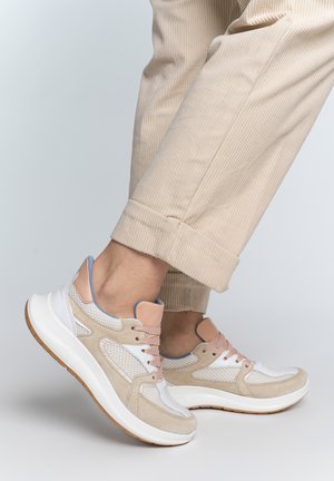 Beige and white sneakers with pink laces worn with beige corduroy pants, showing one foot lifted on a light gray background.