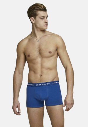 Blue cotton boxer shorts with a black elastic waistband featuring "JACK & JONES" logo. Model with short hair stands sideways, showing torso.