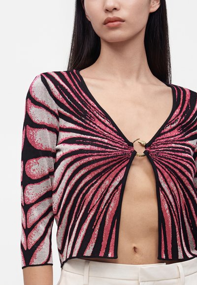 Femme portant un haut à motif abstrait noir et rose avec des manches trois-quarts, fixé par un anneau doré décoratif sur la poitrine.