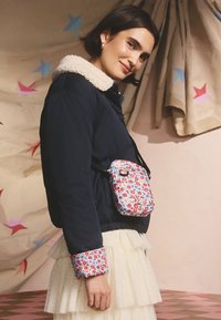 Kvinna som bär en marinblå jacka med blommiga muddarskärmar, en liten blommig axelremsväska och en lager-på-lager krämfärgad kjol, ståendes framför en bakgrund med stjärnmönster.