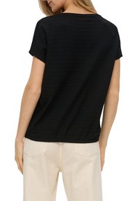 Žena v jednobarevném černém tričku s krátkým rukávem a světle béžových kalhotách, zachycena zezadu na bílém pozadí.