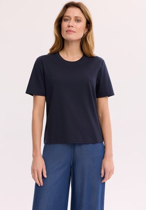 Kobieta stojąca, ubrana w gładki ciemnogranatowy T-shirt z krótkim rękawem oraz luźne niebieskie dżinsy na jasnym tle.