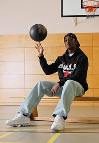 Felpa nera con logo "Chicago Bulls", jeans azzurri e scarpe da ginnastica bianche. Una palla da basket è bilanciata su una mano. Sfondo di una palestra in legno.