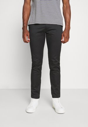 Man wearing black slim-fit pants, white high-top sneakers, and a black-and-white striped t-shirt standing against a plain background.