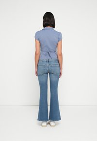 Φωτεινό γαλάζιο μπλουζάκι με κοντά μανίκια και δετή λεπτομέρεια στο πίσω μέρος, συνδυασμένο με καμπάνες μπλε τζιν και λευκά αθλητικά παπούτσια, από την πίσω όψη.