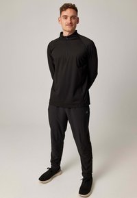 Schwarzes Sportoutfit bestehend aus einem langärmligen Oberteil mit Reißverschluss und einer Hose. Die Textur ist glatt, mit einem figurbetonten Design und minimalem Branding.