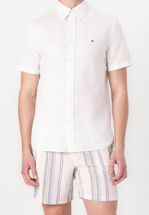 Mand iført en hvid kortærmet skjorte og lyse shorts med lodrette blå, beige og røde striber.