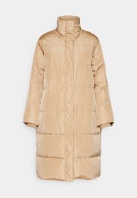 Long manteau matelassé beige avec col montant, fermeture avant cachée, matelassage horizontal et poignets côtelés sur un fond uni.