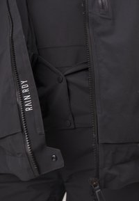 Schwarze wasserdichte Jacke mit einem Reißverschluss vorne, Druckknöpfen und Taschen. Besitzt eine glatte Textur und verstärkte Nähte für Langlebigkeit.