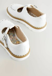 Chaussures en cuir blanc avec un bout arrondi, une sangle avec une boucle et une semelle intérieure à motifs. La semelle extérieure est blanche avec une subtile touche de brun.