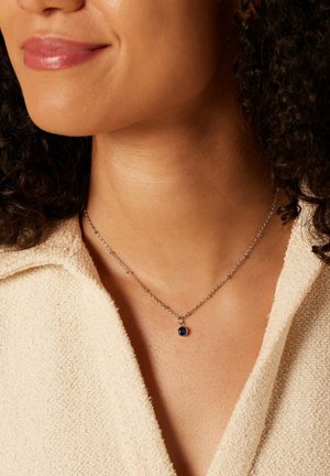 Femme portant une chemise crème texturée et un collier en chaîne argentée avec un pendentif en pierre précieuse bleu foncé.