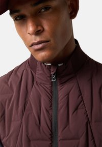 Dunkelrote Steppjacke mit hohem Kragen, ausgestattet mit einem Frontreißverschluss und strukturiertem Stoff. Nahaufnahme des Halses und des oberen Brustbereichs.